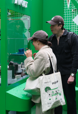 Two people inspecting AVOLT charging accessories in a green pop‑up display; tote bag with AVOLT illustration